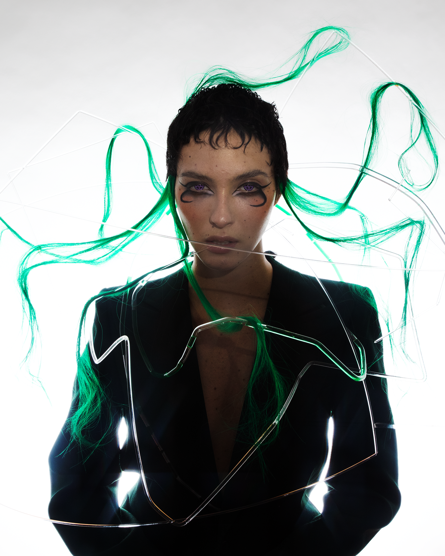 Artistic portrait of a model styled as ‘Medusa,’ featuring vibrant green hair extensions integrated into a structured wire framework. The model wears a sleek black blazer and striking avant-garde makeup with black swirling eyeliner. Styled and directed by Taiga Sato for Infringe Magazine.