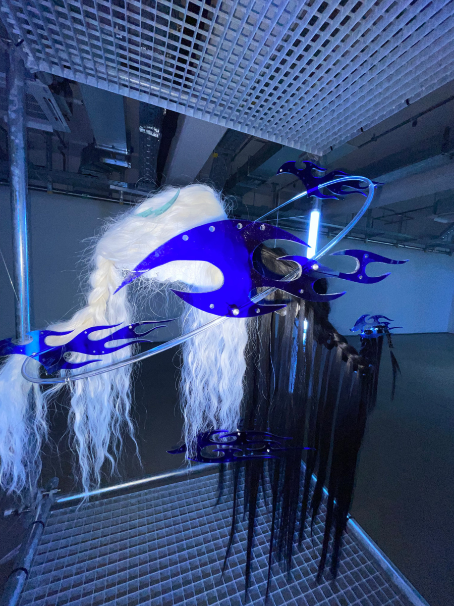 Taiga Sato’s hair sculpture ‘KIRIN,’ showcasing braided and flowing strands of black and white hair intertwined with striking blue metallic flame-like elements in a futuristic gallery setting.