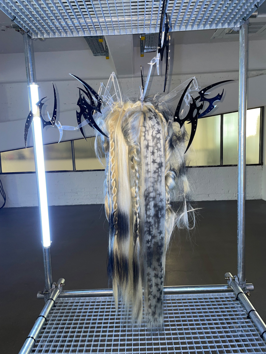 Taiga Sato’s hair sculpture ‘HACHI,’ featuring intricate braids, stenciled star patterns, and flowing black and blonde strands, adorned with sharp metallic blade-like accents in a contemporary exhibition setting.