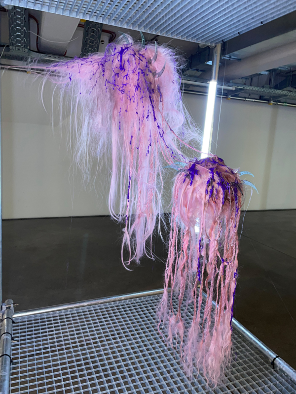 Two sculptural hair installations by Taiga Sato, featuring cascading pink and purple synthetic hair with intricate textures, displayed in an industrial-style gallery setting.