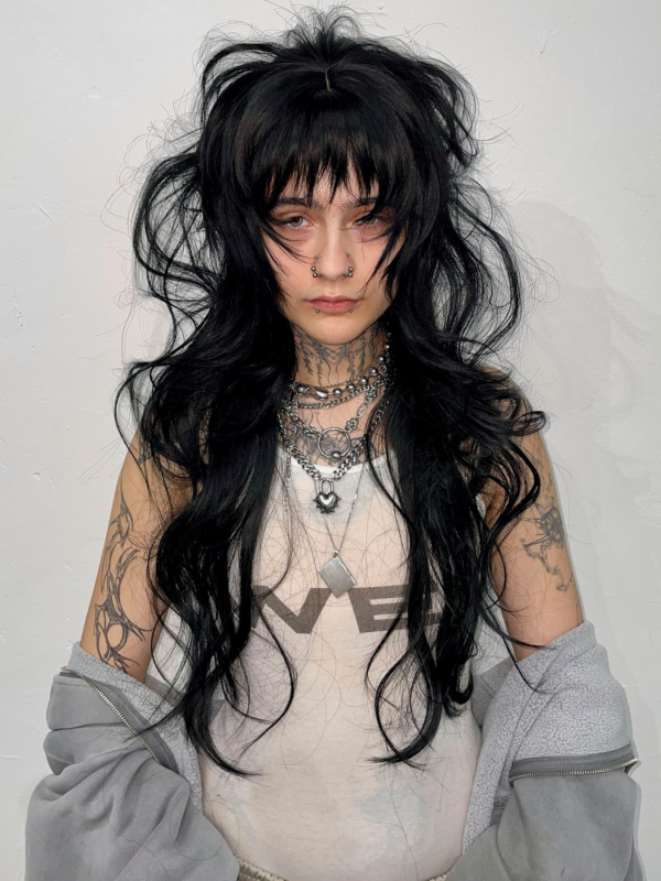 Kolja Tinkova wearing a striking black wig styled by Taiga Sato, featuring voluminous layers, sharp bangs, and a bold, edgy aesthetic, complemented by tattoos, piercings, and layered necklaces.