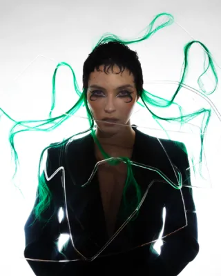 Artistic portrait of a model styled as ‘Medusa,’ featuring vibrant green hair extensions integrated into a structured wire framework. The model wears a sleek black blazer and striking avant-garde makeup with black swirling eyeliner. Styled and directed by Taiga Sato for Infringe Magazine.